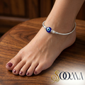 Ankle bracelet with a blue eye charm on a foot against a wooden background, featuring the brand 'Sooma'.