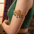 Close up profile view of a model wearing a gold plated antique temple jewelry armlet with red and green gemstone drops against a green silk dress