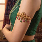 Close up profile view of a model wearing a gold plated antique temple jewelry armlet with red and green gemstone drops against a green silk dress