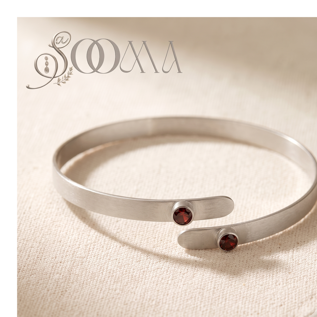 A silver-toned open cuff bracelet with two small round dark red gemstone accents, displayed on a textured beige fabric background. The brand name 'SOAA' is visible in the top left corner.