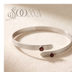 A silver-toned open cuff bracelet with two small round dark red gemstone accents, displayed on a textured beige fabric background. The brand name 'SOAA' is visible in the top left corner.