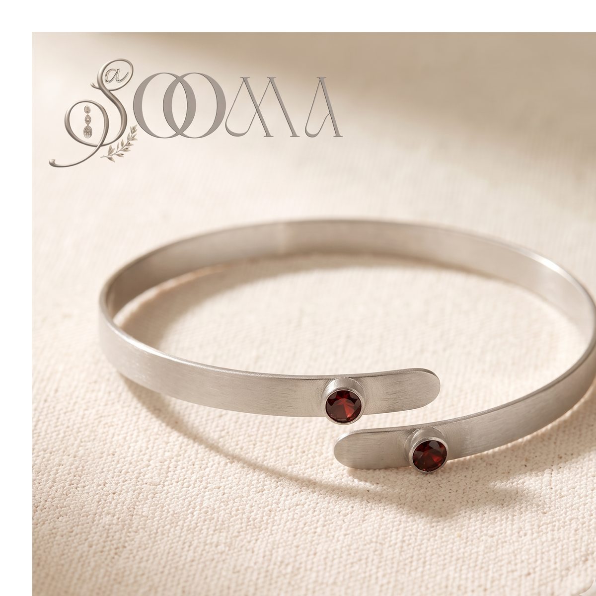 A silver-toned open cuff bracelet with two small round dark red gemstone accents, displayed on a textured beige fabric background. The brand name 'SOAA' is visible in the top left corner.