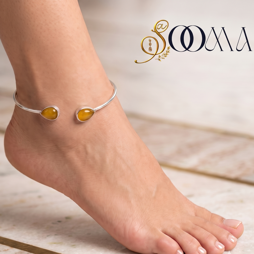 A close-up of a person's ankle and foot wearing a silver-toned bangle anklet with two oval amber-yellow gemstones, set against a light marble floor. The brand name 'SOOMA' is visible in the top right corner.