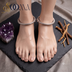 A pair of silver-toned snake-skin ankle bracelets worn on a model's feet, styled on a dark slate board with a purple amethyst crystal and dried roots. The brand logo 'SOOMA' is visible in the top left corner.