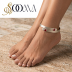 A silver-toned wrap-style ankle bracelet with two small red gemstone accents, shown worn on a person's ankle with bare feet on a beige stone surface. The SOOMA brand logo appears in the top left corner.