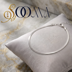 A silver snake-chain anklet displayed on a light gray satin pillow, set against a marble background with a gold and navy SOOMA logo in the top left corner.