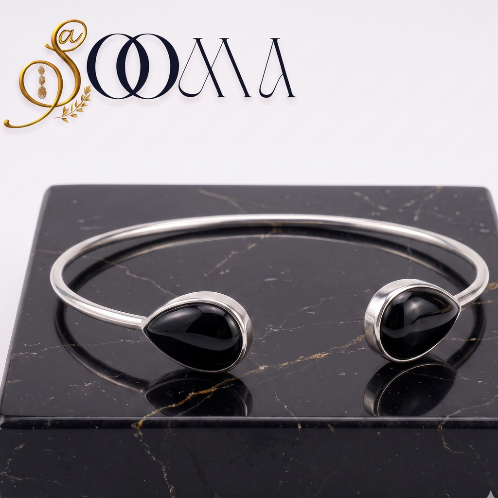 A silver cuff bracelet with two black teardrop gemstone settings, displayed on a black marble block with the SOOMA brand logo in the top left corner.