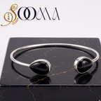 A silver cuff bracelet with two black teardrop gemstone settings, displayed on a black marble block with the SOOMA brand logo in the top left corner.