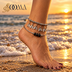 A close-up of a person's foot and ankle wearing two anklets outdoors on a stone path with greenery and flowers. The anklets are silver-toned, one with small leaf charms and the other with black beads.