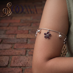 A silver-toned bangle bracelet with a central purple flower charm and small pearl-like beads, shown worn on a person's upper arm against a brick wall background. The brand name 'SOANA' is visible in the top left corner.