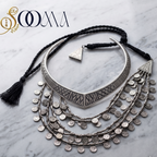 A silver-toned bohemian-style collar necklace with geometric patterns, layered dangling coin chains, and black braided cord with tassels, displayed on a white marble background with a 'SOOMA' brand logo in the top left corner.