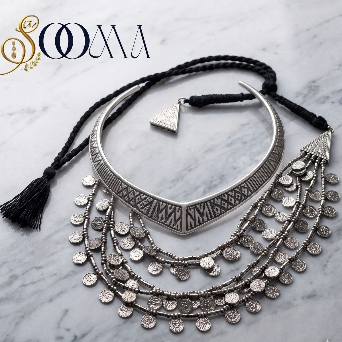 A silver-toned bohemian-style collar necklace with geometric patterns, layered dangling coin chains, and black braided cord with tassels, displayed on a white marble background with a 'SOOMA' brand logo in the top left corner.