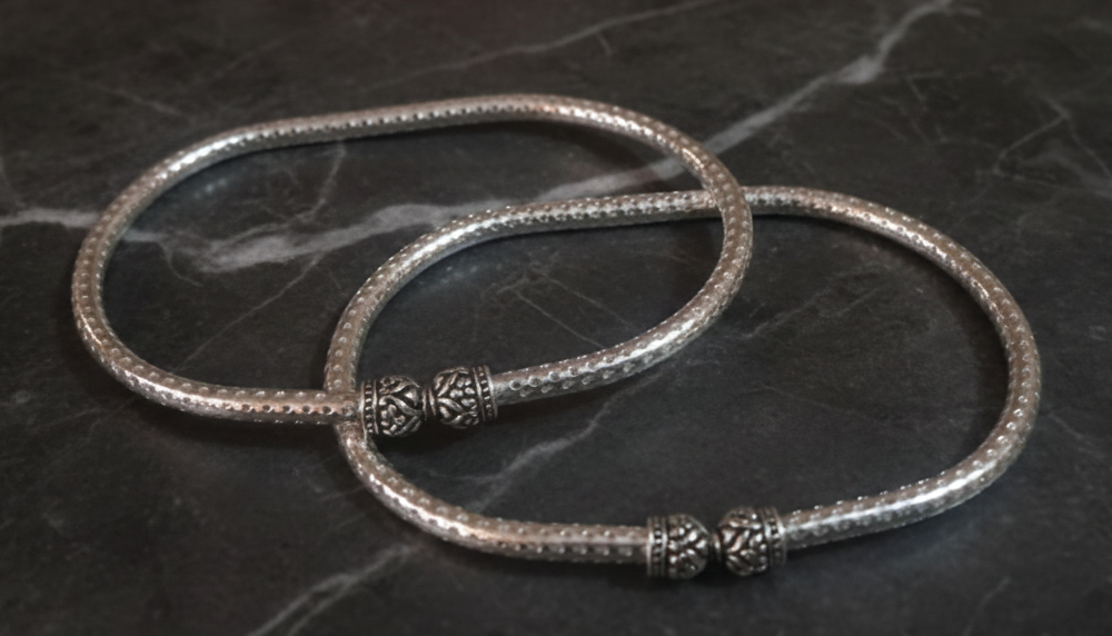 Two silver ankle bracelets with decorative elements on a dark marble surface