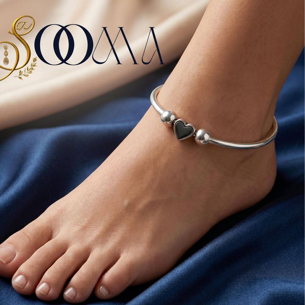 Silver anklet with heart charm on a foot against a blue fabric background, featuring the brand 'Sooma'.