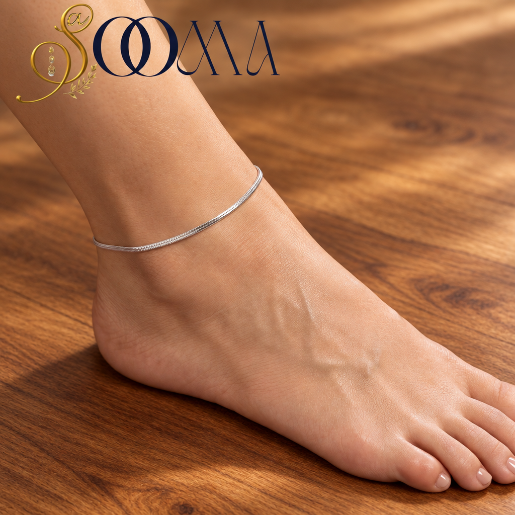 A close-up of a person's foot wearing a thin silver-toned ankle bracelet, resting on a purple velvet cushion. The brand logo 'SOOMA' is visible in the top left corner.