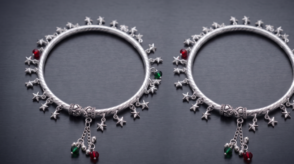 Two silver ankle kadas with star and gemstone charms on a dark background
