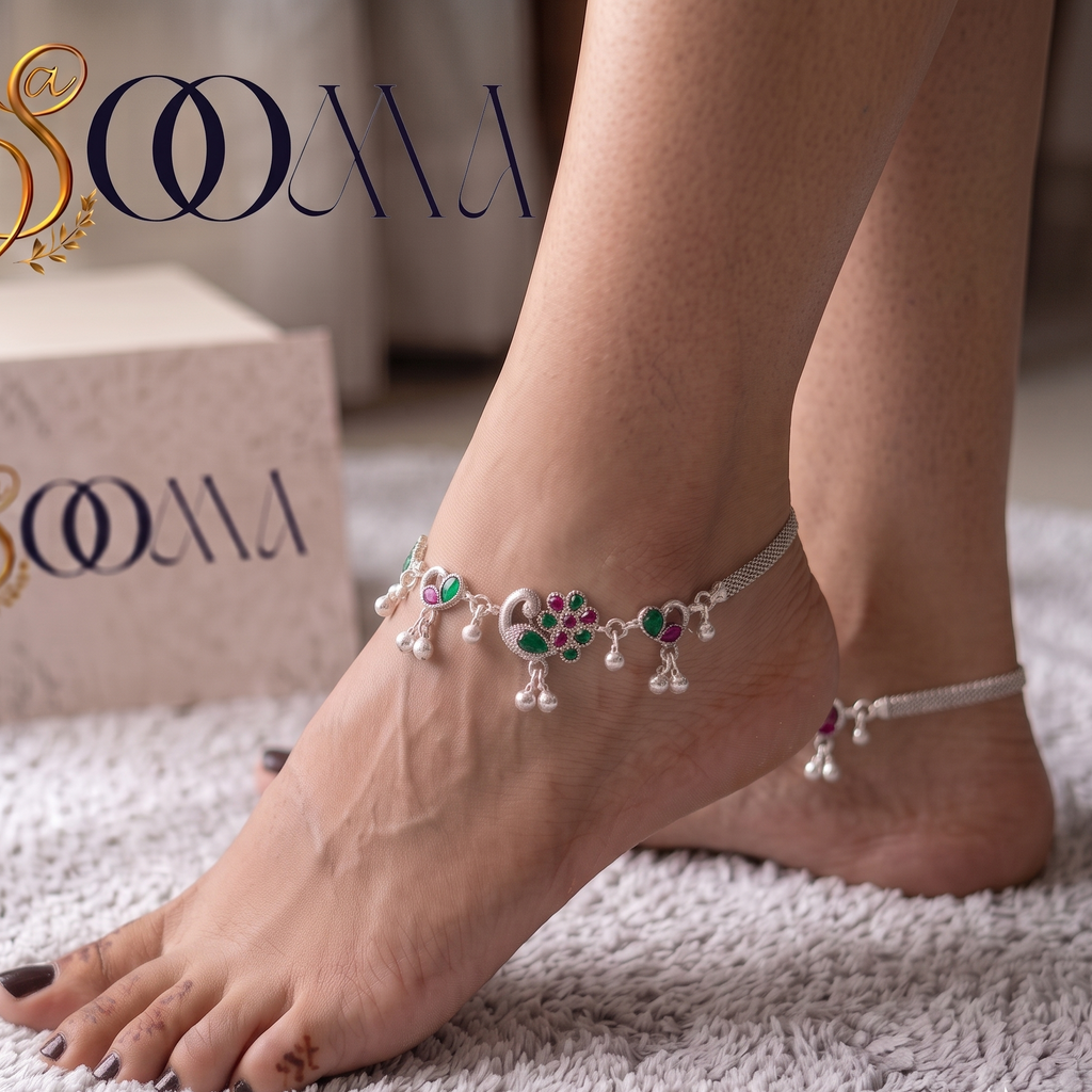A pair of bare feet wearing delicate silver anklets with colorful floral and dangling bead details, standing on a white textured rug. A branded SOOMA box is visible in the background.