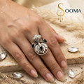 A silver-toned ring with a detailed floral and crown-like design, worn on a hand. The ring features black teardrop-shaped stones and a central black oval stone.