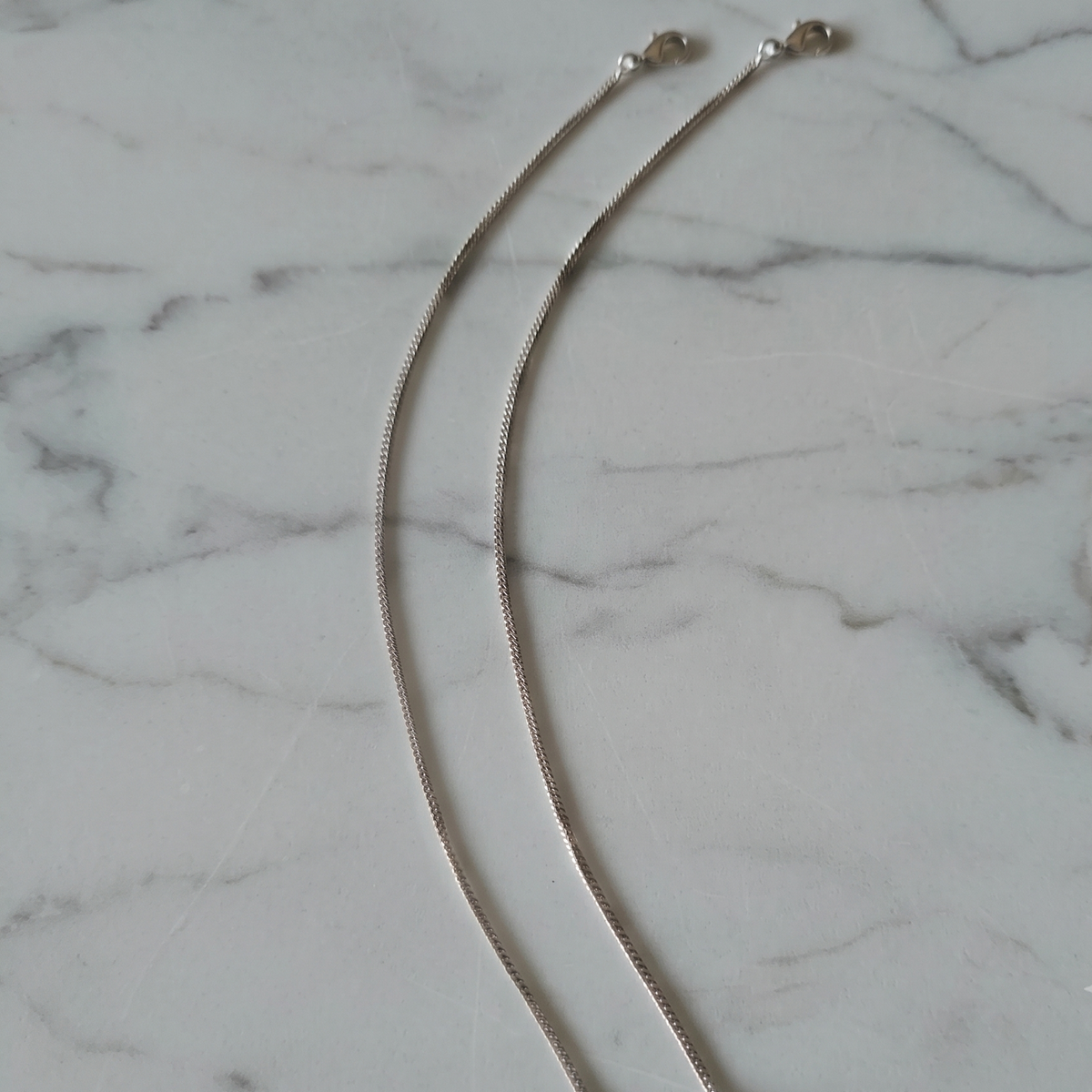 Two silver chain anklets with lobster clasps, laid out on a white marble surface.