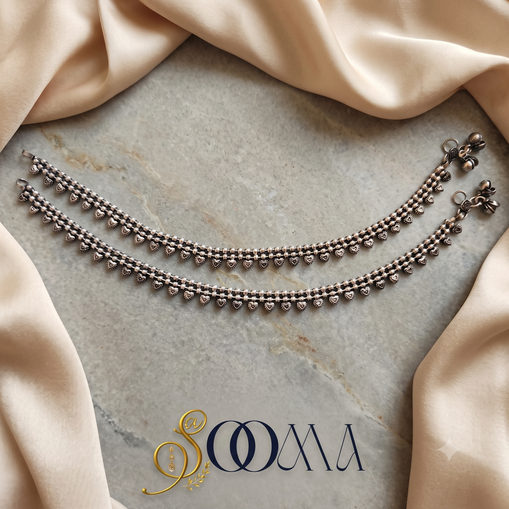 Pair of handcrafted oxidized silver double-strand anklets with heart drops displayed flat on marble and cream silk fabric