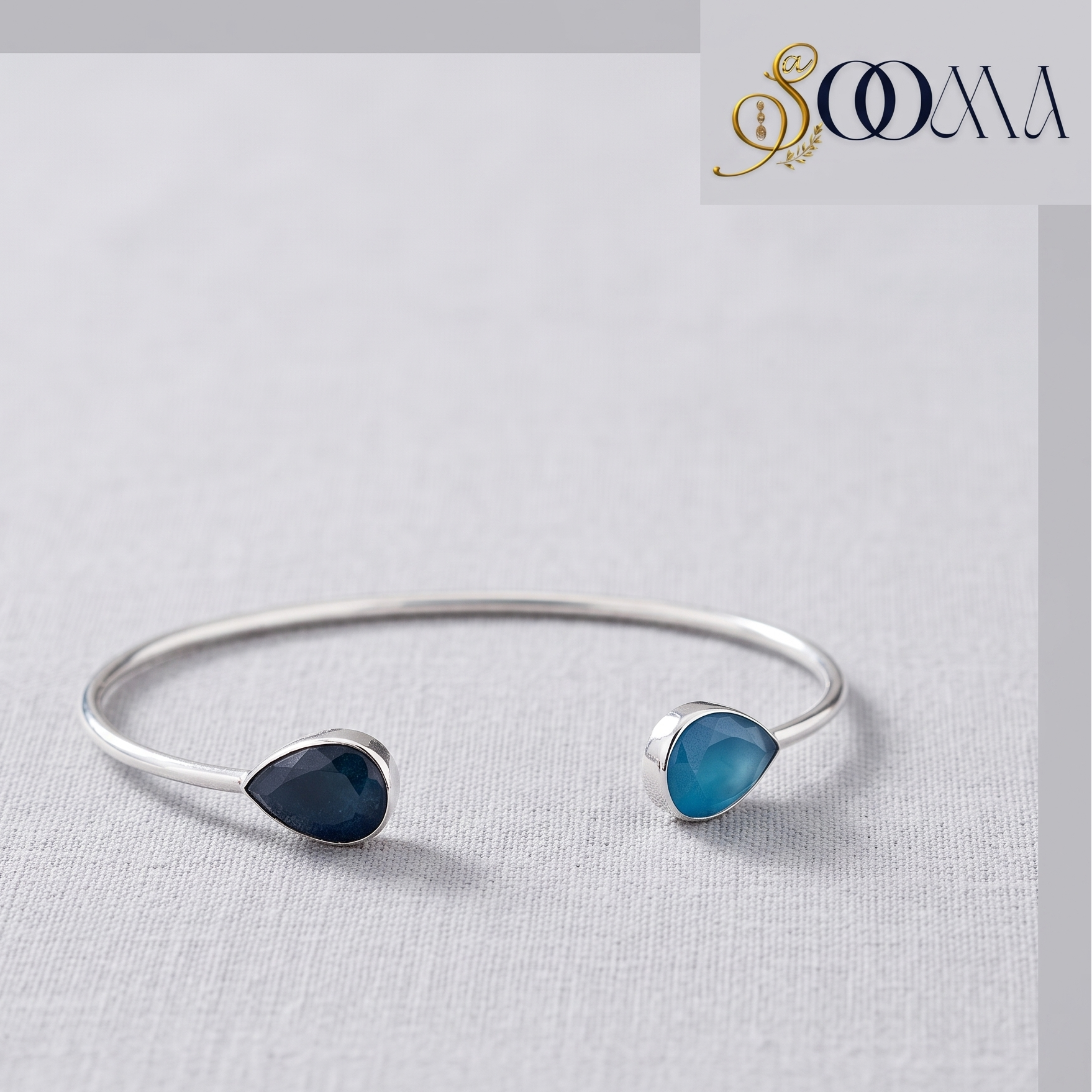 A silver open anklebracelet with two teardrop-shaped gemstones, one dark navy blue and one light blue, displayed on a textured light gray fabric background. The brand logo 'OMMA' is visible in the top right corner.