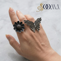 Hand wearing two rings, one floral and one butterfly-shaped, on a light background with 'Sooma' branding.