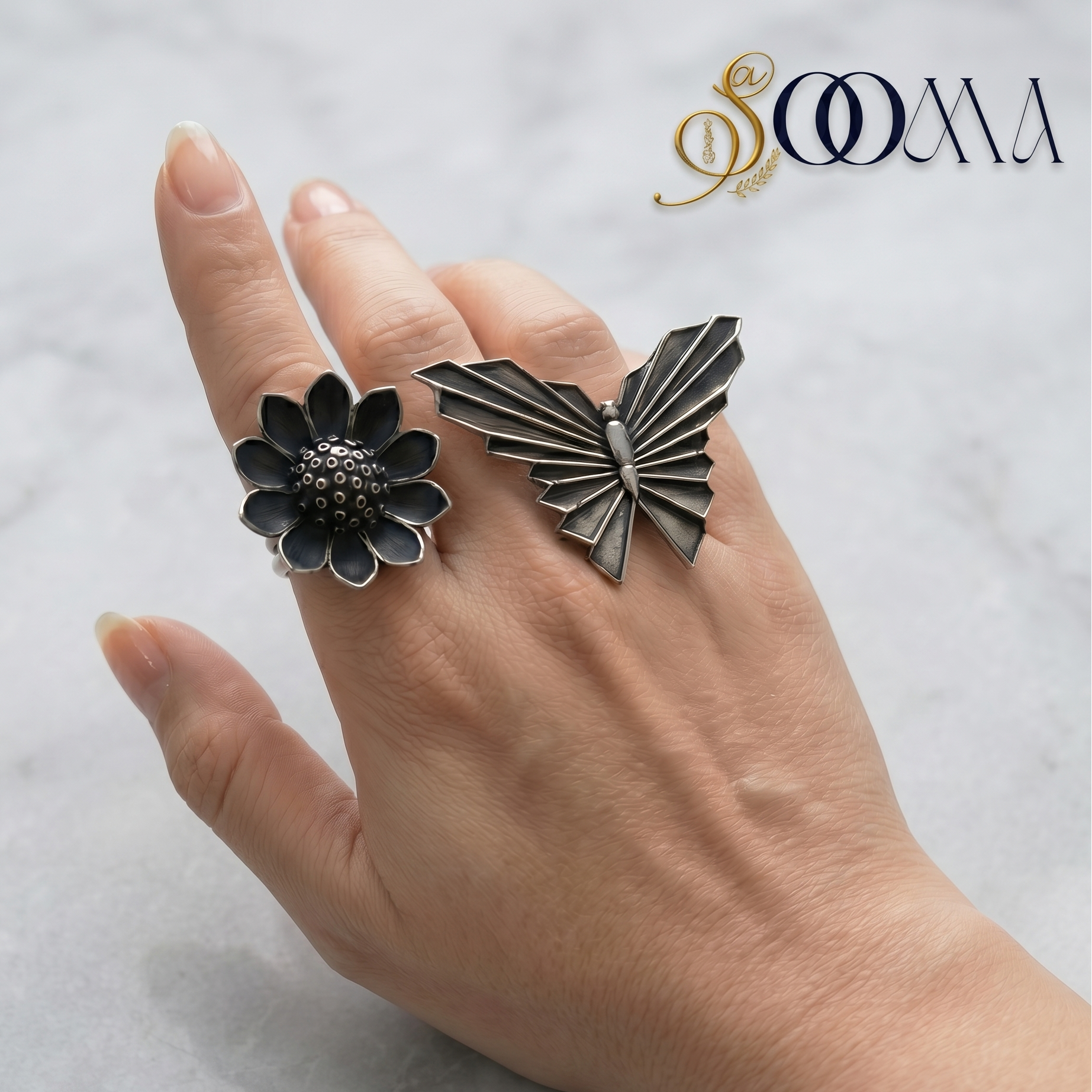 Hand wearing two rings, one floral and one butterfly-shaped, on a light background with 'Sooma' branding.