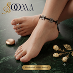 A close-up lifestyle shot of a person's feet and ankles wearing a dark brown braided anklet with silver butterfly charms, along with matching silver toe rings, set against a dark green marble background with dried flowers and a brand logo 'SOOMA' at the top.