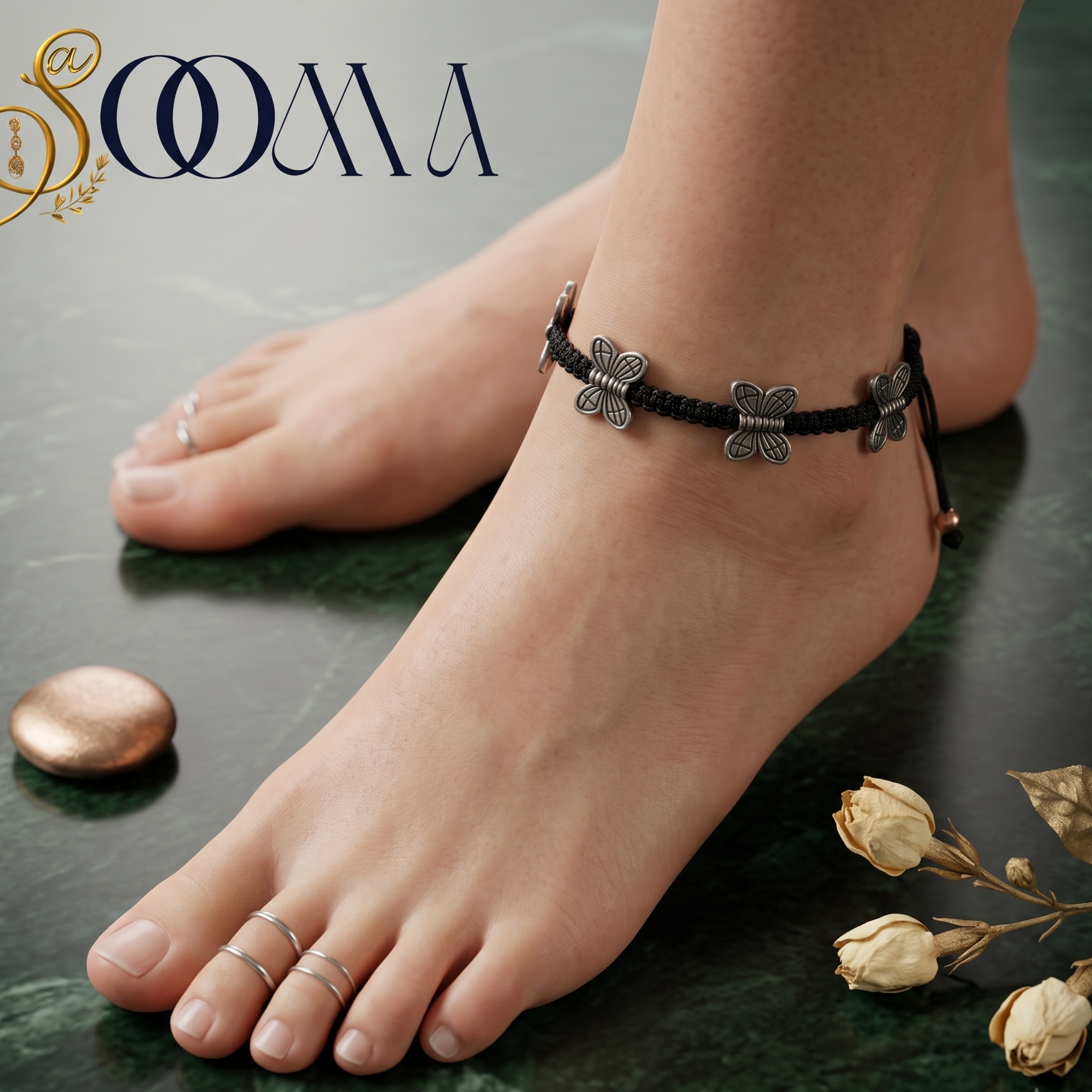 A close-up lifestyle shot of a person's feet and ankles wearing a black braided anklet with silver butterfly charms. The background is a dark green marble surface with decorative stones and dried flowers, and the brand logo 'SOOMA' is visible in the top left corner.