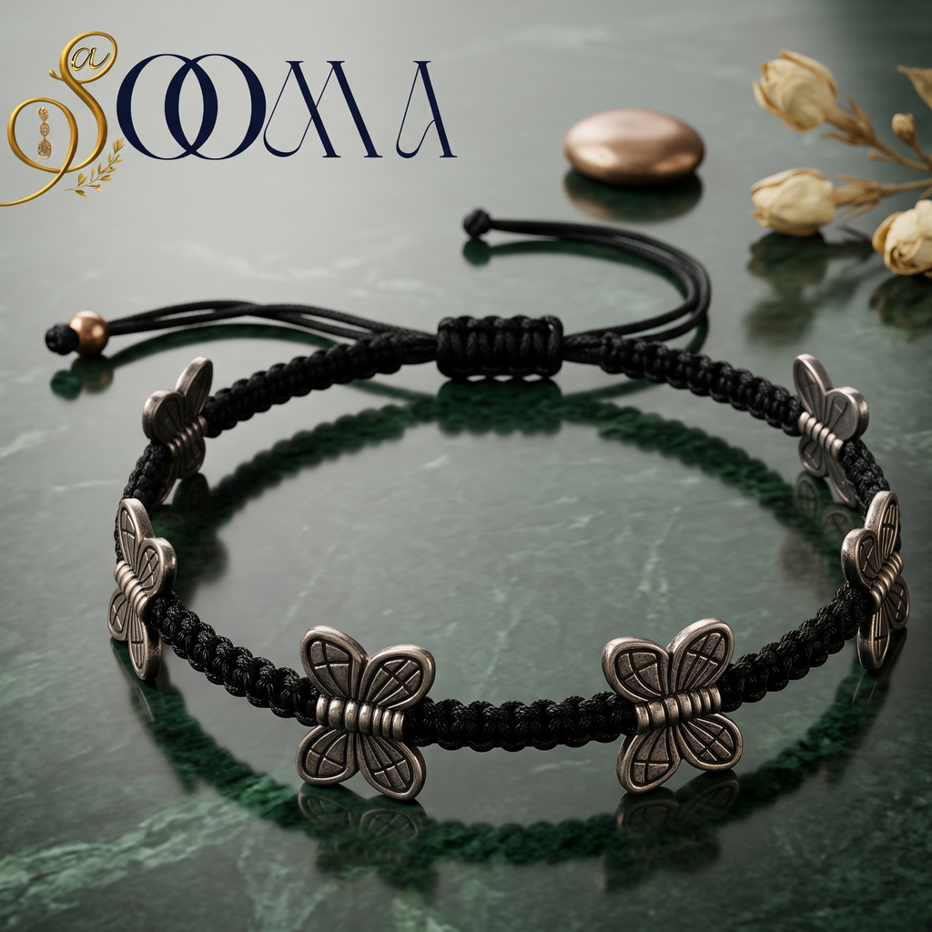 A black braided cord anklet with four silver butterfly charms, displayed on a dark green marble surface with decorative dried flowers and a brand logo 'SOOMA' in the top left.