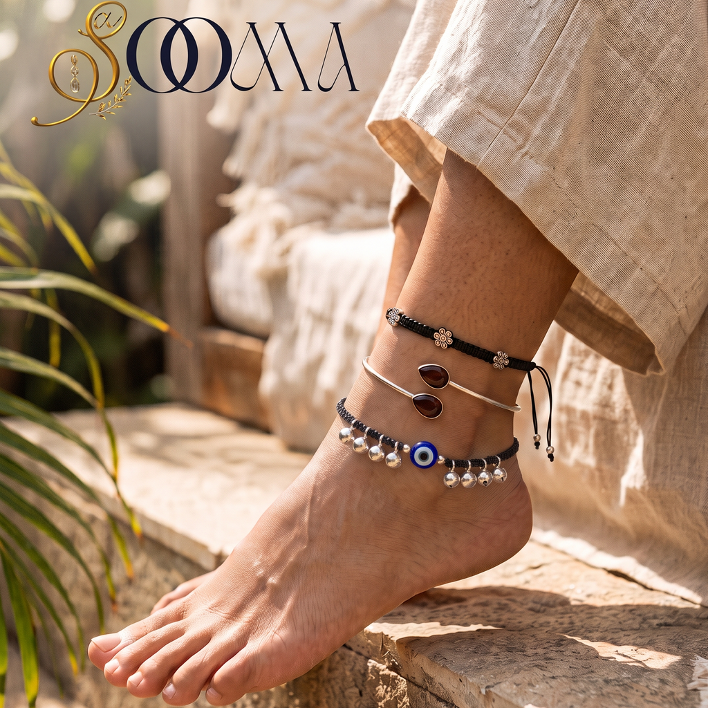 Side profile view of model seated outdoors wearing a 3-piece bohemian silver ankle stack with Evil Eye charm and black stone bypass cuff