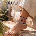 Side profile view of model seated outdoors wearing a 3-piece bohemian silver ankle stack with Evil Eye charm and black stone bypass cuff