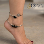 A close-up of a person's ankle and foot wearing two delicate silver anklets. One anklet features a small rose charm, and the other has a row of glossy black beads.