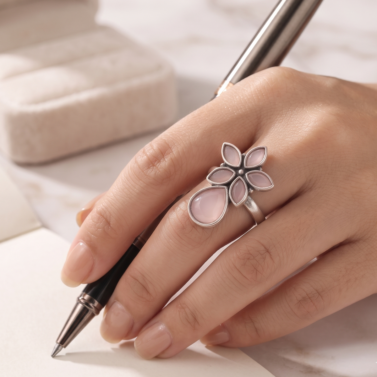 A silver flower ring with pale pink teardrop-shaped stones is worn on a person's hand, which is holding a black and silver pen over a white marble surface.