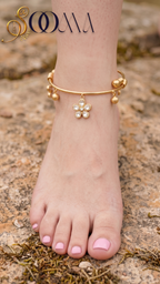 A gold anklet with a dangling flower charm and small bead accents is shown worn on a person's ankle, photographed outdoors on a rocky surface. The brand name SOOMA appears in the upper left corner.