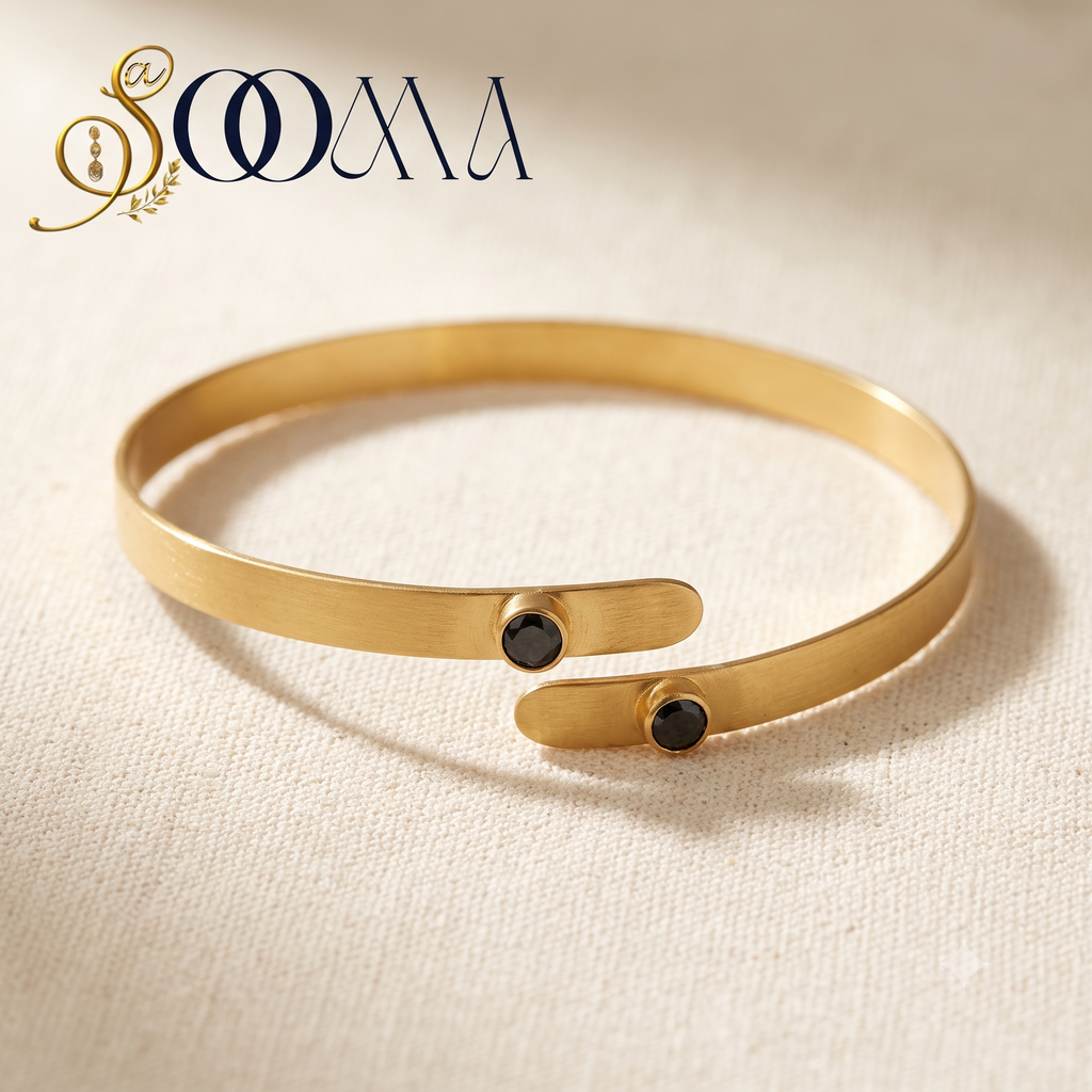 A gold open cuff bracelet with two small black oval stones, displayed on a textured beige fabric background with a brand logo 'SOOMA' in the top left corner.