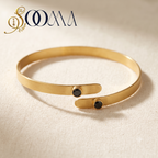 A gold open cuff bracelet with two small black oval stones, displayed on a textured beige fabric background with a brand logo 'SOOMA' in the top left corner.