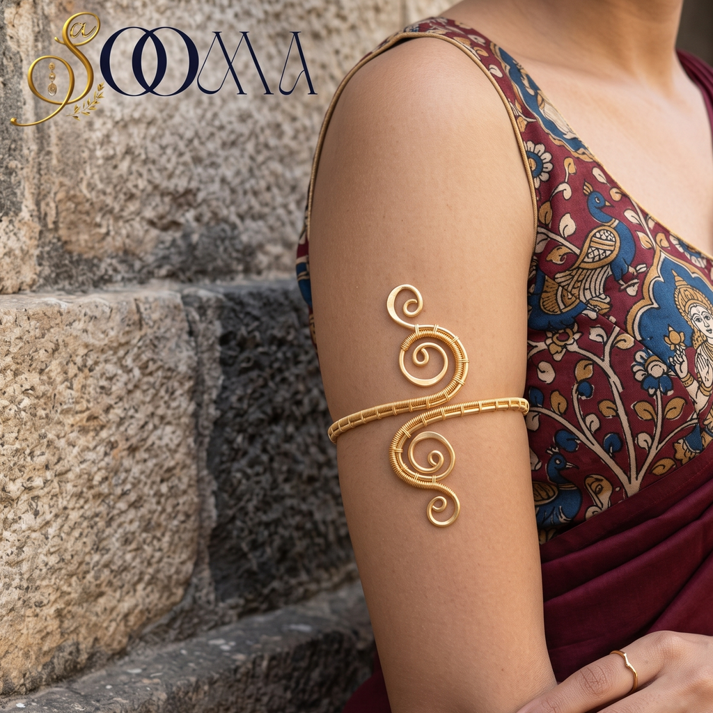 A woman wearing a gold-toned decorative bangle with a swirling, coiled design on her upper arm, paired with a patterned blouse and draped maroon saree, standing against a stone wall. The brand name 'SOUMA' is visible in the top left corner.