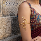 A woman wearing a gold-toned decorative bangle with a swirling, coiled design on her upper arm, paired with a patterned blouse and draped maroon saree, standing against a stone wall. The brand name 'SOUMA' is visible in the top left corner.