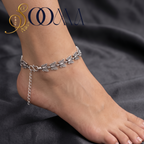 A silver-toned anklet with detailed leaf and vine motifs worn on a person's ankle, shown against a dark gray draped fabric background. The brand name 'SOOMA' is visible in the top left corner.