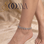 A silver-toned leaf-patterned anklet worn on a woman's ankle, shown against a beige patterned fabric background with the brand logo 'SOOMA' in the top left corner.