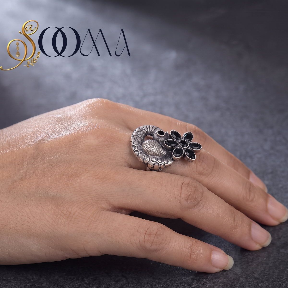 A silver peacock ring with black enamel details and a flower accent, worn on a person's finger against a dark gray textured background. The brand logo 'SOOMA' is visible in the top left corner.