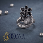 A silver-toned ring shaped like a peacock with a black flower on its back, photographed on a dark textured surface with small scattered beads around it. The brand name 'SoOMA' appears in the lower left corner.