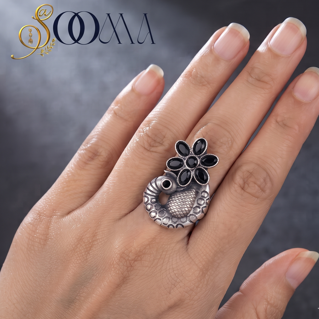A hand wearing a silver ring with a peacock and flower motif, featuring black inlay details. The brand logo 'SOOMA' is visible in the top left corner against a dark textured background.
