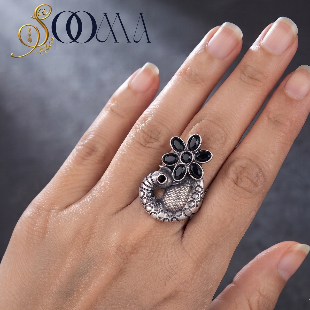 A hand wearing a silver ring with a peacock and flower motif, featuring black inlay details. The brand logo 'SOOMA' is visible in the top left corner against a dark textured background.