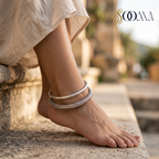Side profile view of model feet seated on stone steps wearing a 2-piece oxidized silver peacock ghungroo HASLI kada and engraved floral ankle stack