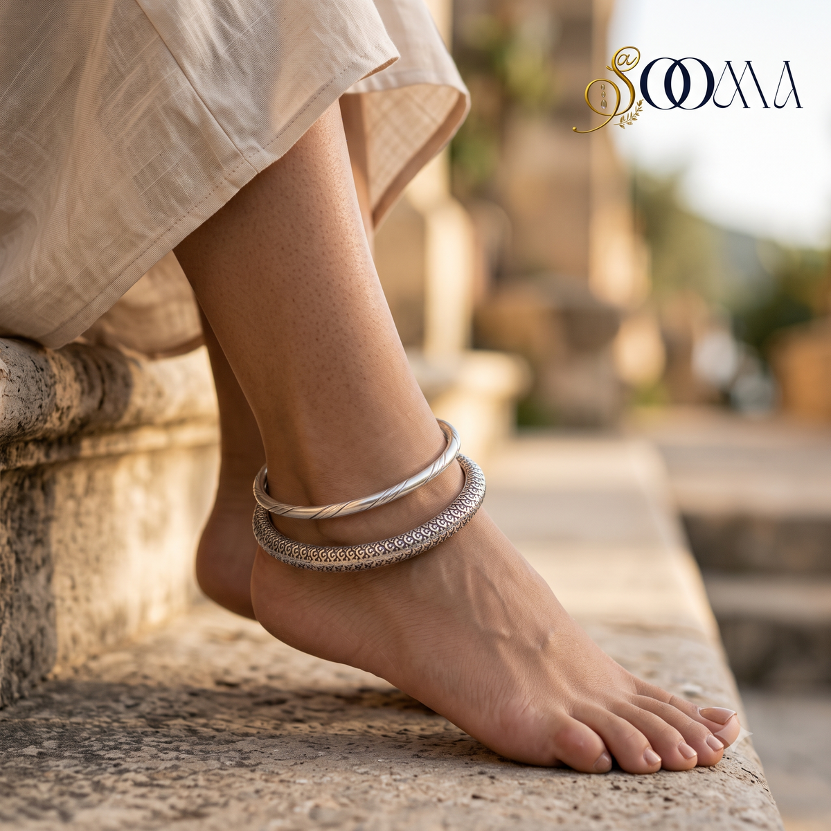 Side profile view of model feet seated on stone steps wearing a 2-piece oxidized silver peacock ghungroo HASLI kada and engraved floral ankle stack