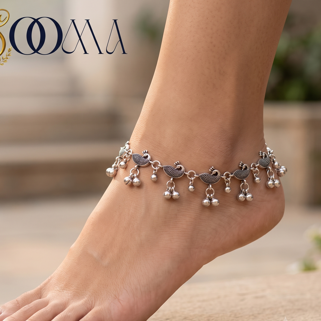 A silver-toned anklet with bird and pearl charms worn on a person's ankle, shown barefoot against a softly blurred outdoor stone background. The brand name 'SOOMA' appears in the top left corner.