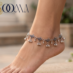 A silver-toned anklet with bird and pearl charms worn on a person's ankle, shown barefoot against a softly blurred outdoor stone background. The brand name 'SOOMA' appears in the top left corner.