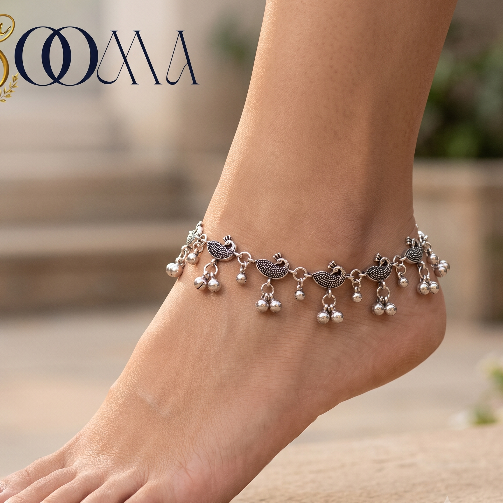 A silver-toned anklet with bird and pearl charms worn on a person's ankle, shown barefoot against a softly blurred outdoor stone background. The brand name 'SOOMA' appears in the top left corner.